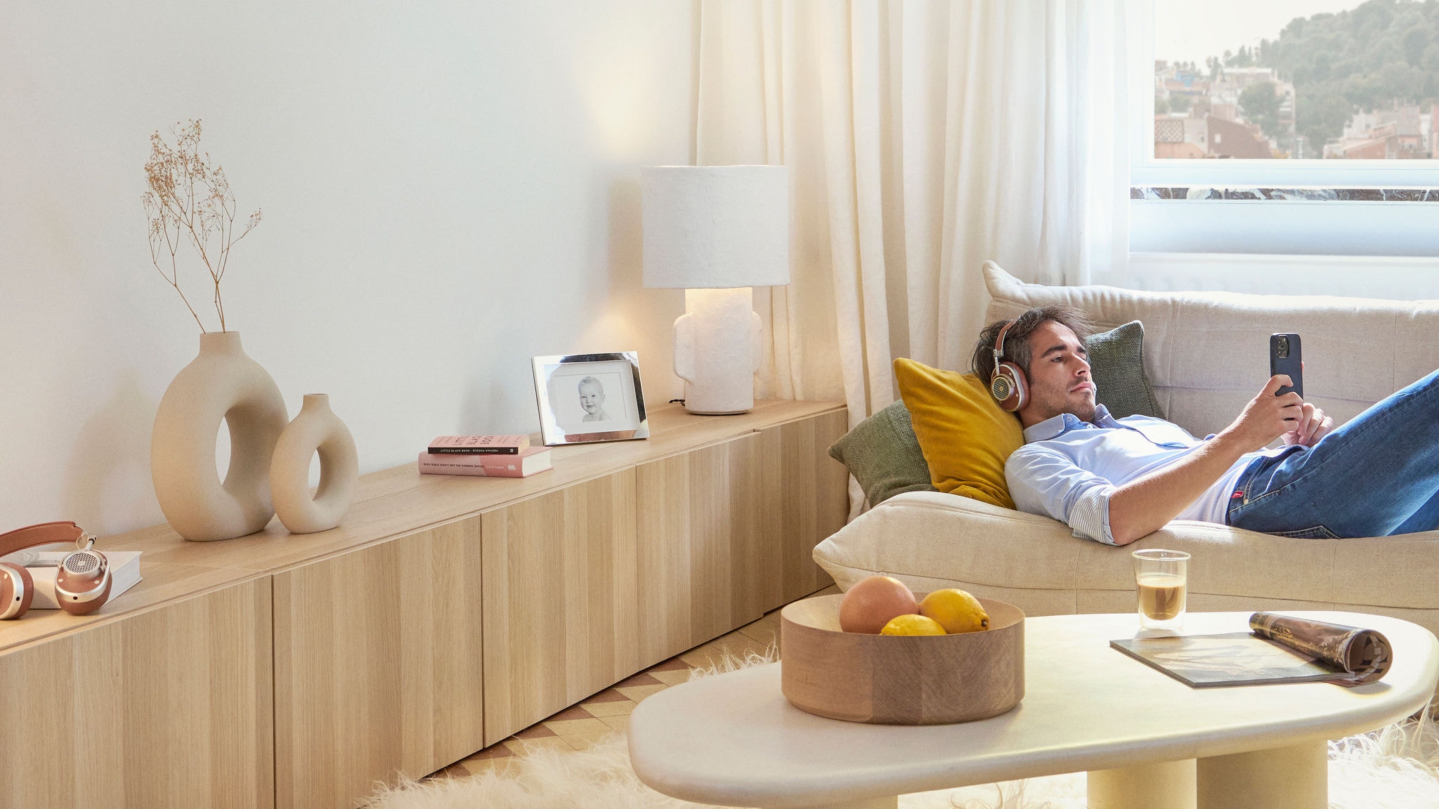 En mand ligger på en sofa i en stue med hovedtelefoner på og scroller på sin telefon.