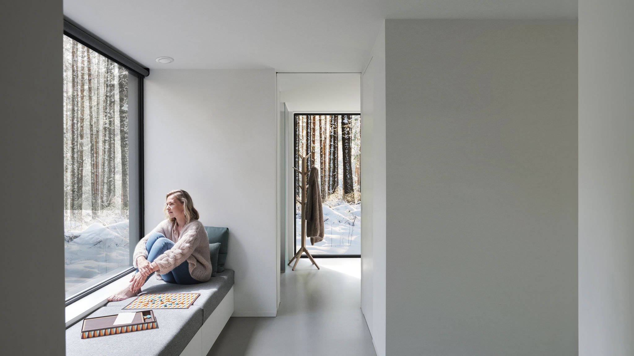 På afstand ses en kvinde, der sidder i bare fødder ved et vindue og kigger ud på træerne og sneen.