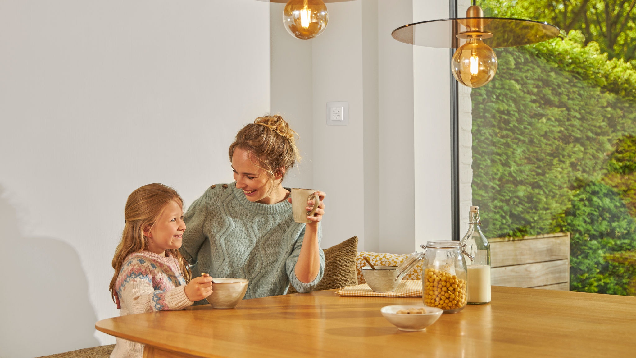 En kvinde og hendes datter spiser morgenmad, mens de nyder den brugervenlige betjening af den smarte ventilationsløsnings CO2- og fugtstyring via fjernbetjeningen