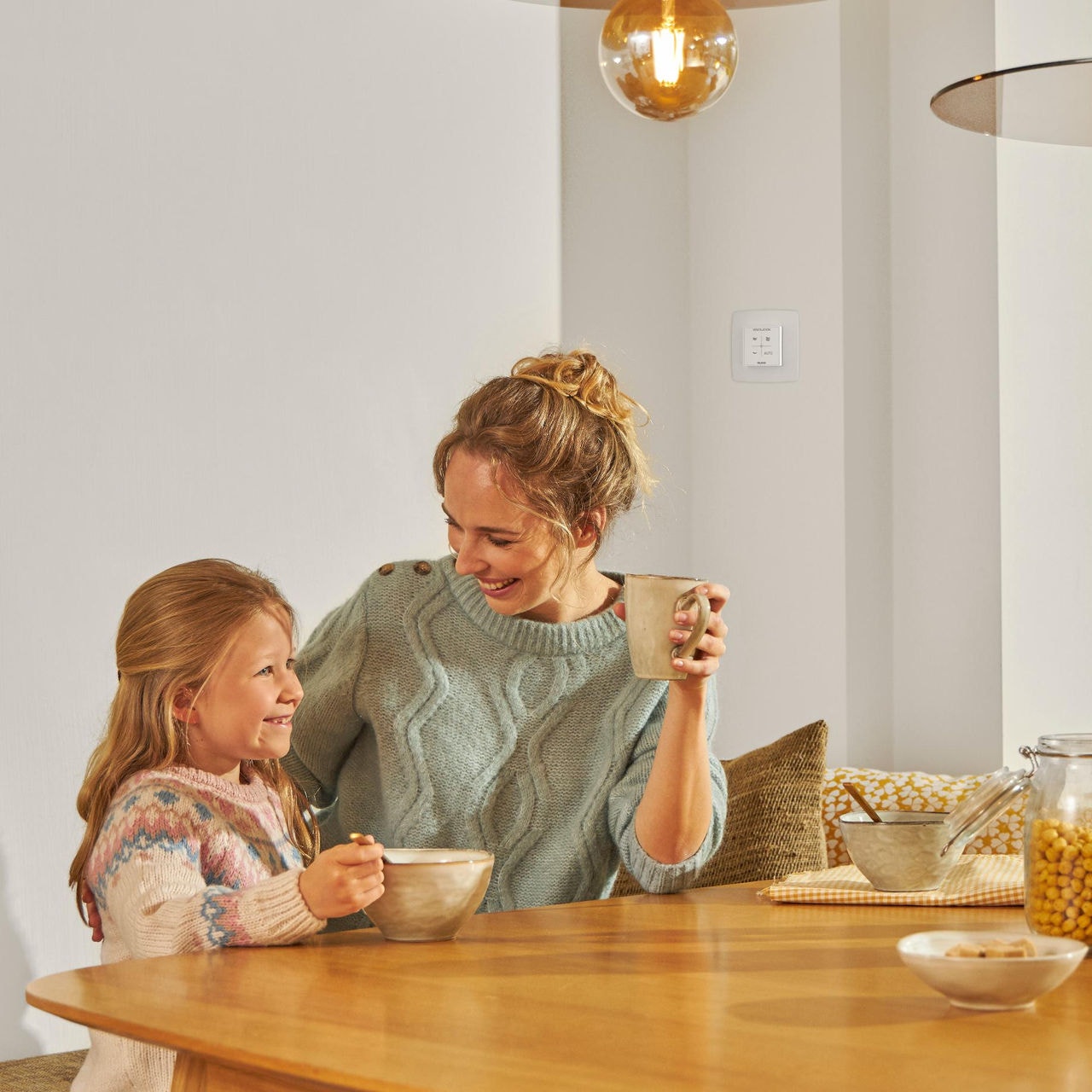  Young girl eating from a bowl seated at a table next to a woman with a mug in hand, both smiling, happy and relaxed, enjoying healthy air.