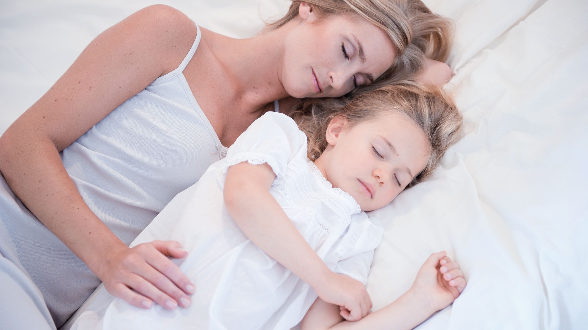 Mor og hendes lille datter sover ved siden af hinanden i en hvid seng i ren luft.