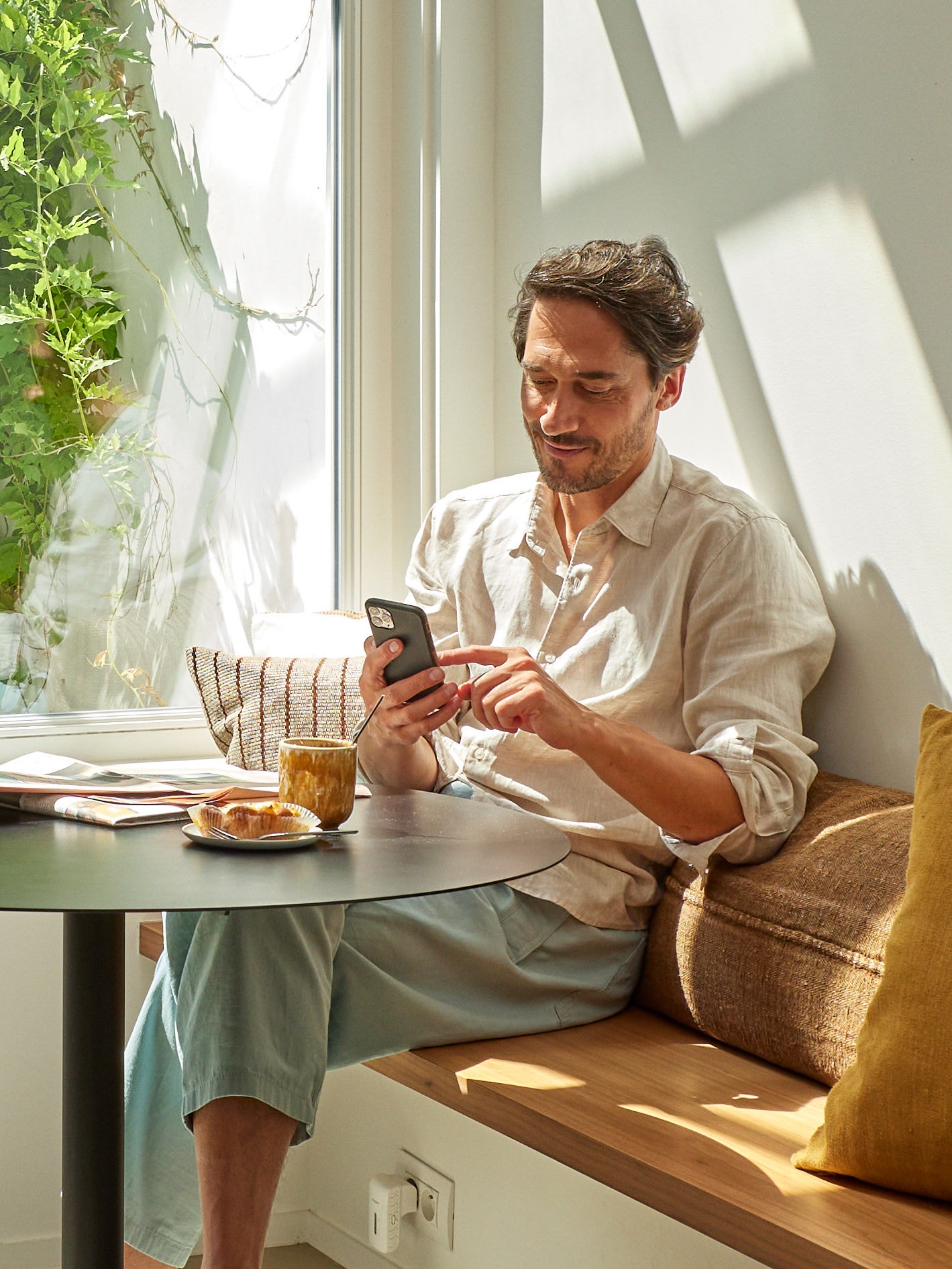 En mand sidder ved et bord og slapper af, mens han kigger på sin telefon med en kop kaffe og noget mad foran sig.