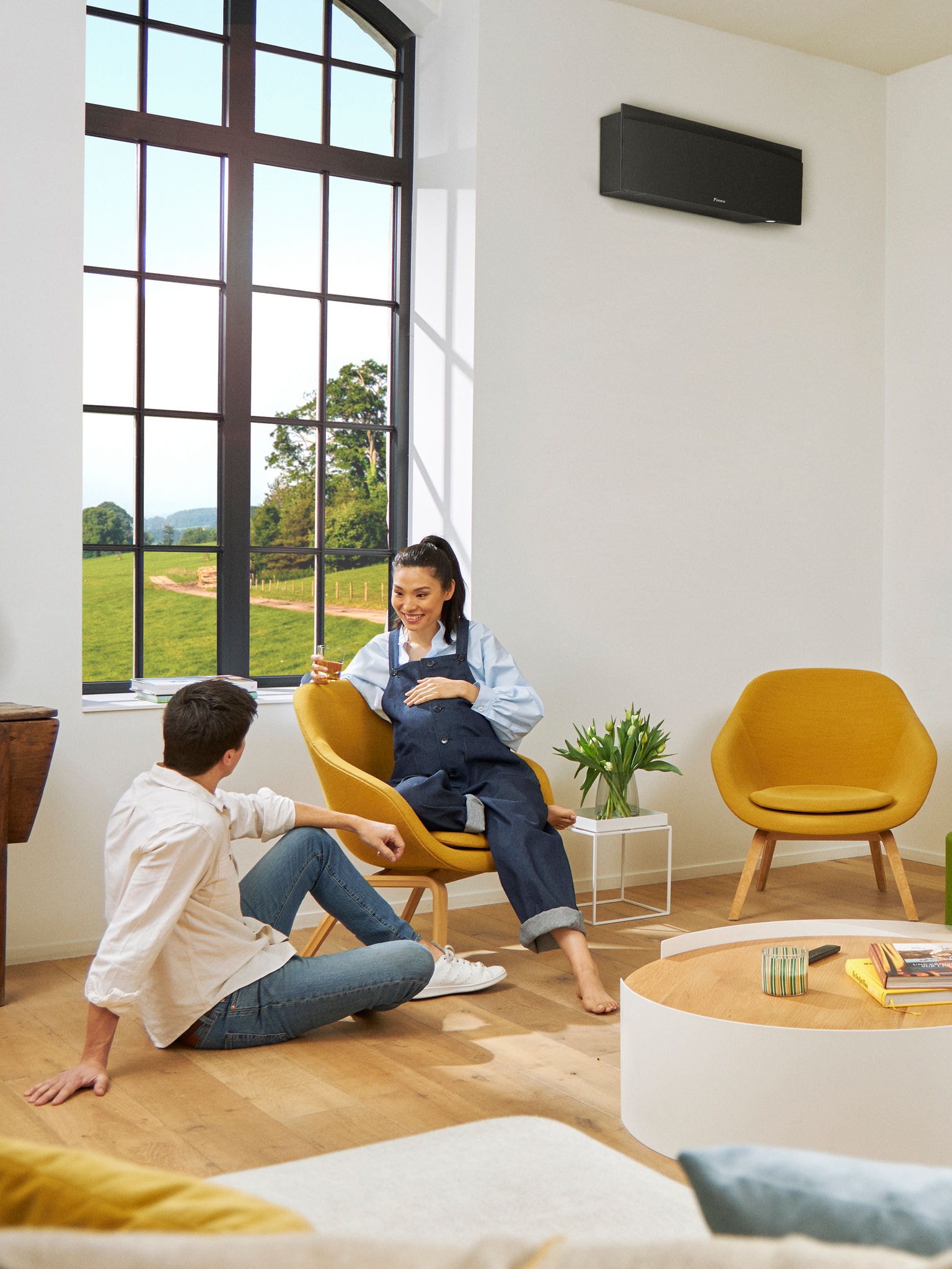 En mand og en kvinde sidder foran et stort vindue i et lyst rum med en sort, vægmonteret varmepumpe i baggrunden