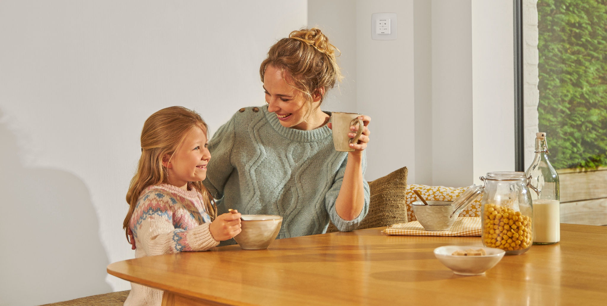 En lille pige sidder ved et bord og spiser ved siden af en kvinde, der holder et krus i hånden, begge smiler og er glade og afslappede, mens de nyder den rene luft.