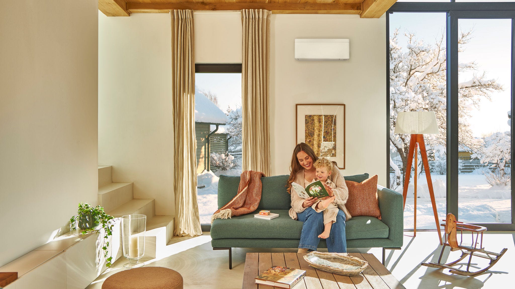 En mor sidder på sofaen under en vægmonteret luft-til-luft varmepumpe og læser en børnebog med sit lille barn på skødet og nyder varmen, mens det sner udenfor.